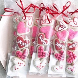 Op de afbeelding: Vier doorzichtige cellofaantassen gevuld met hartvormige suikerkoekjes versierd met roze, rode en witte glazuur. De koekjes hebben verschillende ontwerpen, waaronder harten, XOXO en een ruitpatroon. De tassen zijn vastgebonden met rood lint.