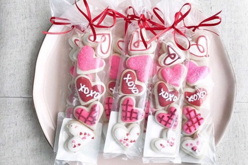 Op de afbeelding: Vier doorzichtige cellofaantassen gevuld met hartvormige suikerkoekjes versierd met roze, rode en witte glazuur. De koekjes hebben verschillende ontwerpen, waaronder harten, XOXO en een ruitpatroon. De tassen zijn vastgebonden met rood lint.