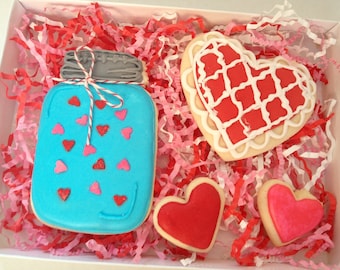 May include: A gift box filled with four decorated sugar cookies. The cookies are shaped like a mason jar, a heart, and two smaller hearts. The mason jar cookie is blue with pink hearts, the large heart cookie is red with white lines, and the smaller hearts are pink. The cookies are surrounded by pink and red shredded paper.