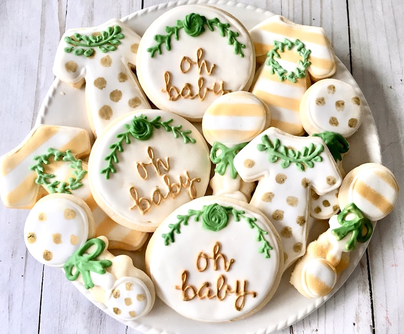 Puede incluir: Un plato de galletas de az&uacute;car decoradas con tem&aacute;tica de baby shower. Las galletas est&aacute;n decoradas con hojas verdes, puntos dorados y la frase "Oh Baby" en letras doradas.