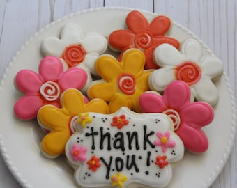 Op de afbeelding: Een bord met gedecoreerde suikerkoekjes in de vorm van bloemen met een bedankje in het midden. De koekjes zijn versierd met roze, gele, oranje en witte glazuur.