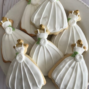 May include: A plate of six white sugar cookies decorated with white icing and gold accents. Each cookie is shaped like a wedding dress.