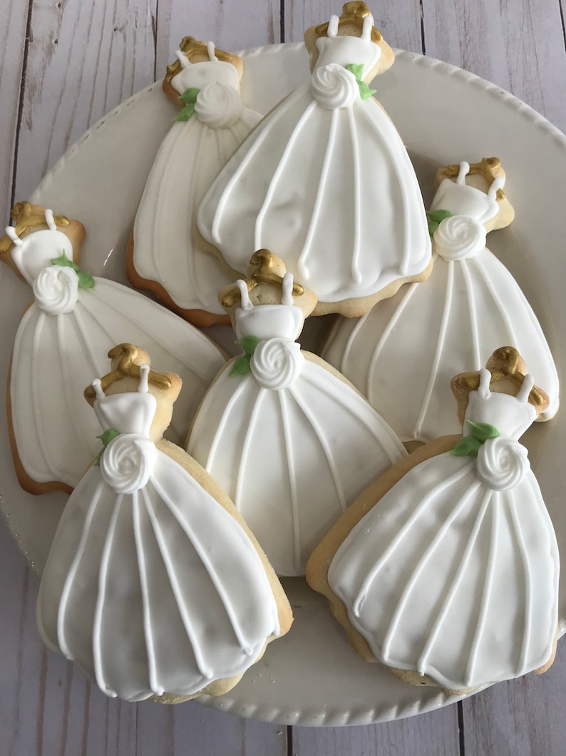 May include: A plate of six white sugar cookies decorated with white icing and gold accents. Each cookie is shaped like a wedding dress.
