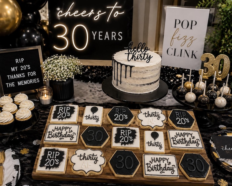 May include: A 30th birthday party display featuring a white cake with black drip icing and a hello thirty topper, surrounded by black and white cookies with text like RIP 20's and Happy Birthday, plus cake pops and signage reading cheers to 30 years.