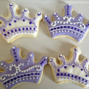 May include: Four purple and white royal crown-shaped sugar cookies decorated with white and purple icing.
