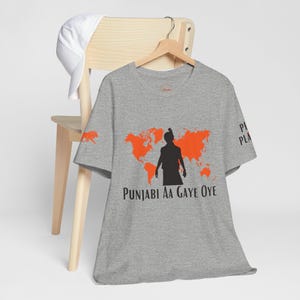 May include: A grey t-shirt featuring an orange world map and a black silhouette figure. The text "PUNJABI AA GAYE OYE" is printed below. A white baseball cap is on a wooden chair.