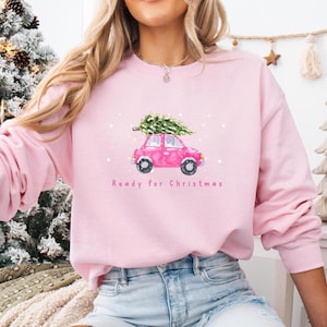 Könnte beinhalten: Ein hellrosa Langarm-Sweatshirt mit einer Grafik eines rosa Autos, das einen grünen Weihnachtsbaum auf dem Dach trägt. Das Design enthält weiße Schneeflocken und den in Rosa unter dem Auto aufgedruckten Text Ready for Christmas.