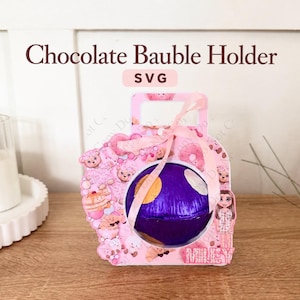 May include: A pink cardboard chocolate bauble holder featuring a circular cutout, a light pink ribbon bow, and a purple foil-wrapped sphere inside. The holder is decorated with cartoon illustrations of bears, donuts, and the name Miley in pink text.