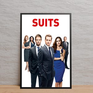 May include: A framed poster featuring the word "SUITS" in red capital letters above a group of people. The group includes men in suits and women in dresses, all standing in front of a white background. The poster is in a black frame.