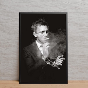 Puede incluir: Impresión enmarcada en blanco y negro de una persona con traje fumando un cigarrillo, rodeada de humo. Una cadena está envuelta alrededor de su muñeca. La impresión se muestra contra una pared gris claro.