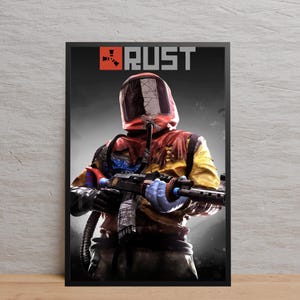 May include: Framed poster with a figure in a hazmat suit holding a firearm. The poster displays the word "RUST" in grey at the top. The figure is clad in a red, yellow, and black suit with a helmet, set against a dark background.