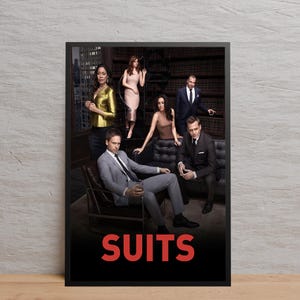 May include: A framed poster featuring the cast of the television show "Suits." The image shows six people in business attire, set against a backdrop of a library and city skyline. The word "SUITS" is displayed in red at the bottom.