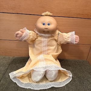 Puede incluir: Una muñeca Cabbage Patch Kid con un vestido amarillo con ribete de encaje blanco. La muñeca tiene el pelo rubio, los ojos azules y una tez rosada.