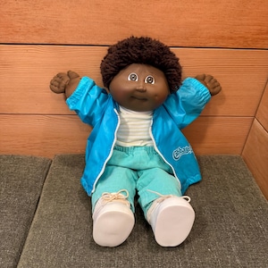 May include: A Cabbage Patch Kid doll with brown skin, brown curly hair, and brown eyes. The doll is wearing a blue jacket, striped shirt, turquoise trousers, and white shoes. The doll is sitting on a grey surface.
