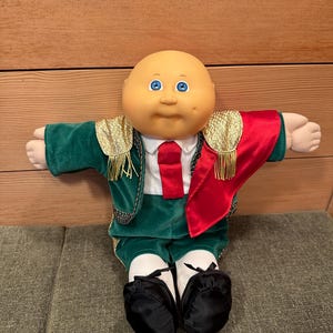 Könnte beinhalten: Cabbage Patch Kid-Puppe in einem Matador-Kostüm. Die Puppe hat einen kahlen Kopf, blaue Augen und einen neutralen Gesichtsausdruck. Das Kostüm ist aus grünem Samt mit goldenen Verzierungen, einem roten Umhang, einem weißen Hemd, einer roten Krawatte und schwarzen Schuhen.