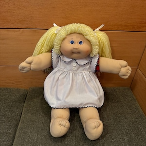 May include: A vintage Cabbage Patch Kid doll with blonde yarn hair in pigtails, blue eyes, and a soft fabric body. The doll is dressed in a white dress with a black scalloped trim and small white buttons.