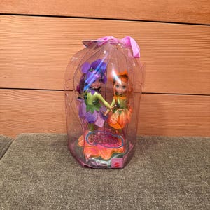 May include: Two fairy dolls in a clear plastic container. One doll has purple hair and a green and purple dress, the other has orange hair and an orange dress. The container is decorated with a pink bow and the word "Fairytopia".