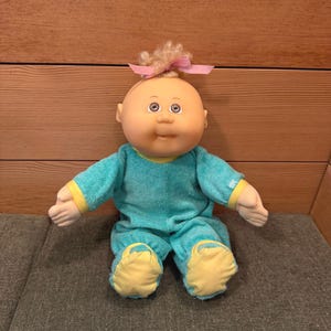 Könnte beinhalten: Eine Cabbage Patch Kids-Puppe mit hellem Teint, blauen Augen und einem kleinen Büschel blondem Haar, das mit einer rosa Schleife gebunden ist. Die Puppe trägt ein türkisfarbenes Frottee-Outfit mit gelben Akzenten an den Manschetten und Füßen. Die Puppe sitzt.