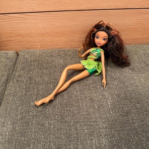 May include: A doll with long brown hair and a green outfit. The doll is lying down on a grey surface. The doll's outfit includes a green top and a skirt. The doll has blue eyes and is wearing a green outfit.