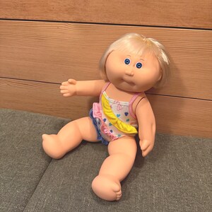 May include: A vintage doll with blonde hair and blue eyes, wearing a swimsuit with a yellow banana-shaped accent. The doll is posed on a grey surface, with its arms and legs extended.