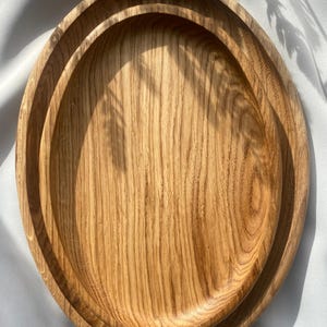 Könnte beinhalten: Zwei ineinander verschachtelte ovale Holztabletts mit einer ausgeprägten natürlichen Maserung in hellbrauner Ausführung. Die Tabletts haben glatte, geschwungene Kanten und ein flaches Beckendesign, das die organische Textur des Holzes hervorhebt.