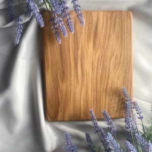 Puede incluir: Una tabla de cortar rectangular de madera con un patrón de veta natural visible, colocada sobre una superficie de tela blanca suave y rodeada de ramas de flores de lavanda moradas que enmarcan las esquinas superior izquierda e inferior derecha.