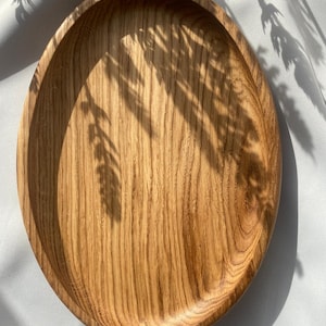 Könnte beinhalten: Ein ovales Holztablett mit einer natürlichen hellbraunen Oberfläche und sichtbarer Holzmaserung. Das Tablett verfügt über eine glatte, flache Vertiefung mit einem erhöhten, abgerundeten Rand, der die organische Textur des Holzes betont.