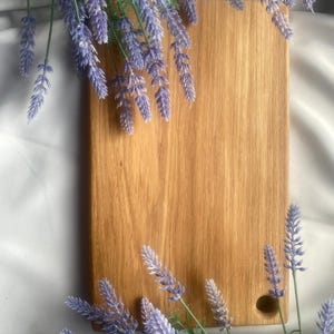 Op de afbeelding: Een rechthoekige houten snijplank met een gladde, lichtbruine nerf en een enkel rond gat nabij de rechterbenedenhoek, omringd door takjes paarse lavendelbloemen.