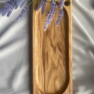 May include: A rectangular wooden tray with a smooth, light brown finish and a prominent natural wood grain pattern. The tray features a deep, elongated oval-shaped carved basin in the center and a small circular hole near the top right corner.