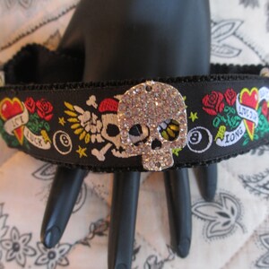 May include: Black choker necklace with a skull charm covered in rhinestones. The choker has a black ribbon with colourful embroidered designs including hearts, roses, a skull and crossbones, and the words "Live Long" and "Ice Luck".