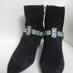 May include: A pair of black suede ankle boots with a decorative buckle and colorful patterned strap. The buckle is silver with a gold accent and the strap is blue, green, and yellow.