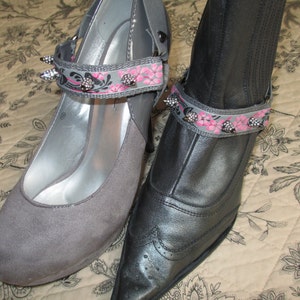 May include: Two pairs of gray shoes with pink and black floral ankle straps. The straps have silver studs and are decorated with pink flowers.