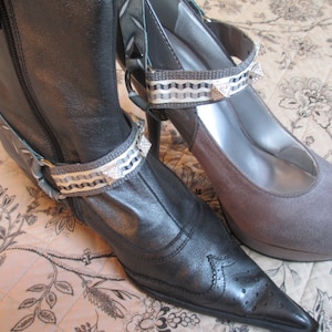 May include: Two pairs of shoes with decorative ankle straps. The straps are made of a white and grey patterned fabric with silver studs. One shoe is a black leather boot with a pointed toe. The other shoe is a grey suede pump with a rounded toe.