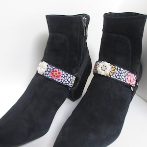 Puede incluir: Un par de botines de ante negro con un adorno floral y de perlas en la correa del tobillo.