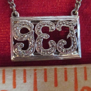 May include: A silver necklace with a rectangular pendant featuring the number 963 in a decorative, ornate font. The numbers are made of small, sparkling stones.