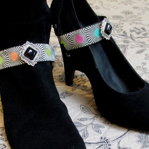 May include: A pair of black suede shoes with a black and white striped ankle strap adorned with colorful circles and a silver buckle with a black gemstone.