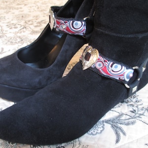May include: A pair of black suede shoes with decorative ankle straps. The straps are made of fabric with a colorful geometric pattern and feature a heart-shaped metal accent.