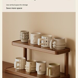May include: A two-tiered wooden shelf holding eight ceramic mugs of various shapes and textures. The text at the top reads: Neat, orderly, and space-saving. Use vertical space for storage. Save more space. The shelf has a dark wood finish.