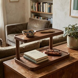 May include: A two-tiered wooden display stand with rounded corners. The top shelf holds a small ceramic bowl with green leaf patterns and a gold pen. The bottom shelf holds two stacked books and a pair of brown-rimmed glasses on a wooden case.