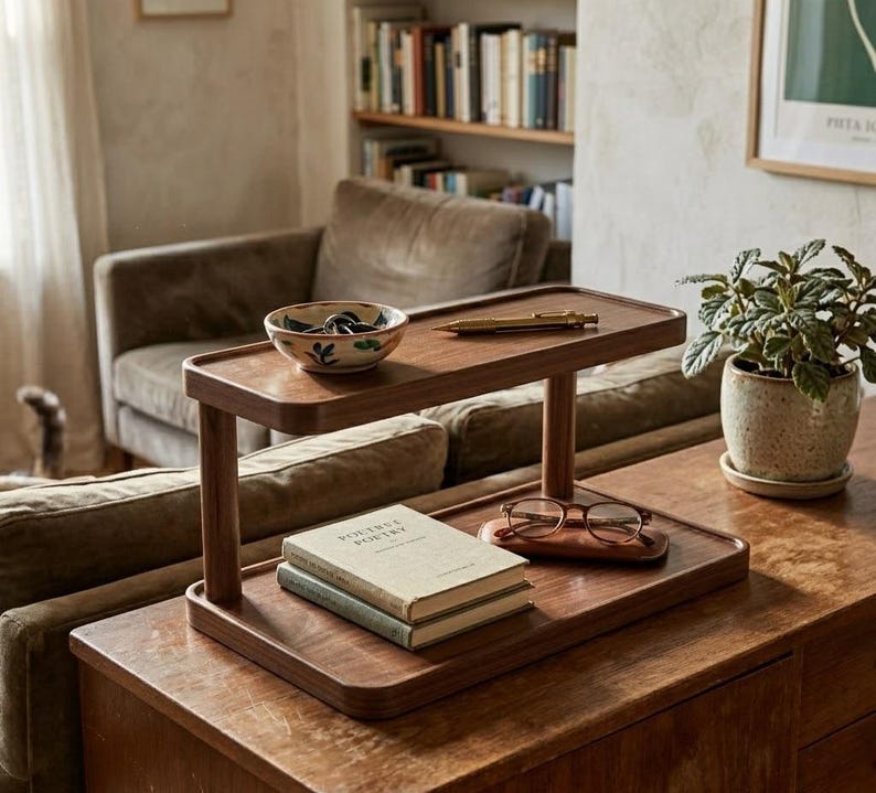 May include: A two-tiered wooden display stand with rounded corners. The top shelf holds a small ceramic bowl with green leaf patterns and a gold pen. The bottom shelf holds two stacked books and a pair of brown-rimmed glasses on a wooden case.