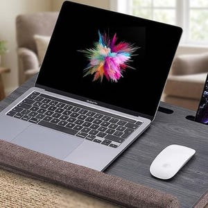 May include: A laptop desk with a grey surface, a built-in phone holder, and a wrist rest. A laptop with a colourful splash screen, a white mouse, and a smartphone are on the desk. The desk is designed for comfort and convenience.