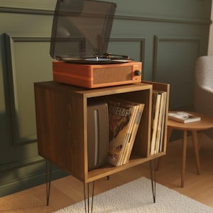 May include: An orange record player with a clear lid sits atop a wooden cabinet with a shelf filled with vinyl records. The cabinet has a dark wood finish and black metal hairpin legs. A white rug is beneath the cabinet.
