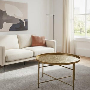 Puede incluir: Una escena de salón con un sofá crema, un cojín naranja y una mesa de centro redonda de latón con un diseño ornamentado. La habitación incluye una alfombra gris y una pintura en la pared.