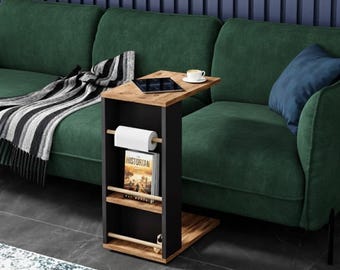 Puede incluir: Una mesa auxiliar moderna con estructura negra y tapa y base de madera. La mesa tiene un revistero incorporado y un portarrollos de papel. Una tableta y una taza de café están sobre la mesa. La mesa está junto a un sofá verde.