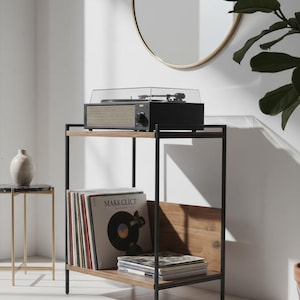 Può includere: Un giradischi nero e beige con una copertura trasparente è posto su una mensola in metallo nero e legno. La mensola contiene dischi in vinile ed è posta contro un muro bianco con uno specchio con cornice dorata e una pianta in vaso.