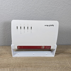 Könnte beinhalten: Weißer Frit!zBox-Router mit roten Akzenten und Kontrollleuchten. Der Router hat den Markennamen oben rechts aufgedruckt. Der Router steht auf einem weißen Ständer und liegt auf einer Holzoberfläche.