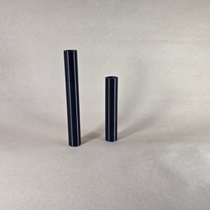 Op de afbeelding: Twee zwarte cilindervormige objecten van verschillende hoogtes staan tegen een neutrale achtergrond. De langste is ongeveer 25 cm hoog, terwijl de kortere ongeveer 13 cm is. Beide hebben een glanzende afwerking.