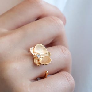 Peut inclure: Une bague dorée en forme de fleur avec une petite pierre précieuse transparente au centre. La bague est portée sur un doigt, et un collier assorti est visible en arrière-plan. Le bijou a une finition mate.