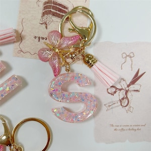 May include: A pink glitter resin letter "S" keychain with a gold clasp, flower charm, and tassel. The letter is filled with iridescent stars and glitter. The keychain has a gold-colored metal clasp.