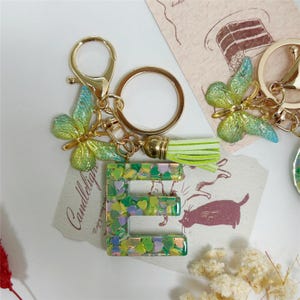 Puede incluir: Un llavero dorado con la letra "E" verde y amarilla llena de confeti en forma de corazón. El llavero presenta un dije de mariposa y una borla verde claro. La palabra "Candlelight" está impresa en una tarjeta pequeña.